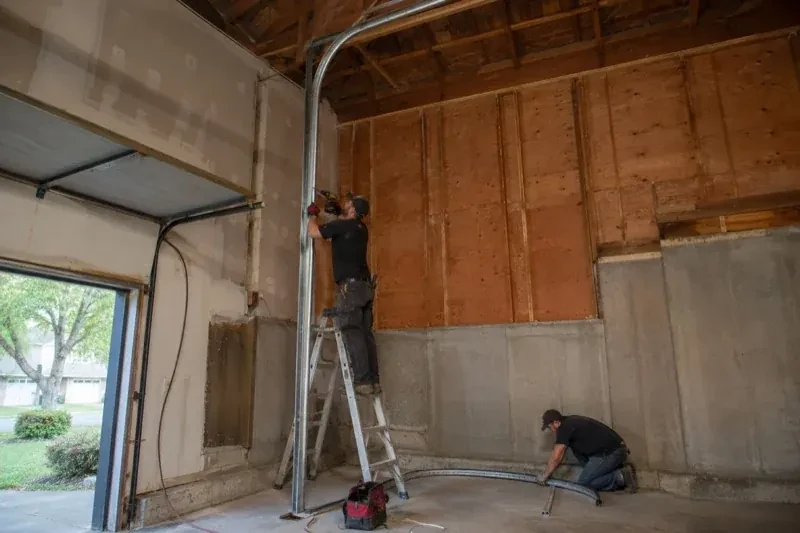 One person on a stepladder working beside a tall curved garage-door track while another kneels beside loose track sections in an unfinished garage. — High-Lift Garage Door Conversion, Georgina
